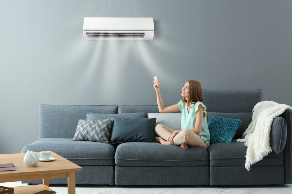 Young Woman Switching On Split System Air Conditioner While Sitting On A Sofa